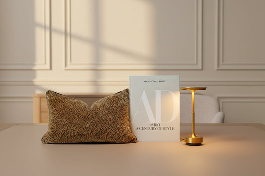 A modern brass table lamp glowing warm yellow, styled on a dark shelf next to a book and a leopard-print pillow. eikhomeco.com