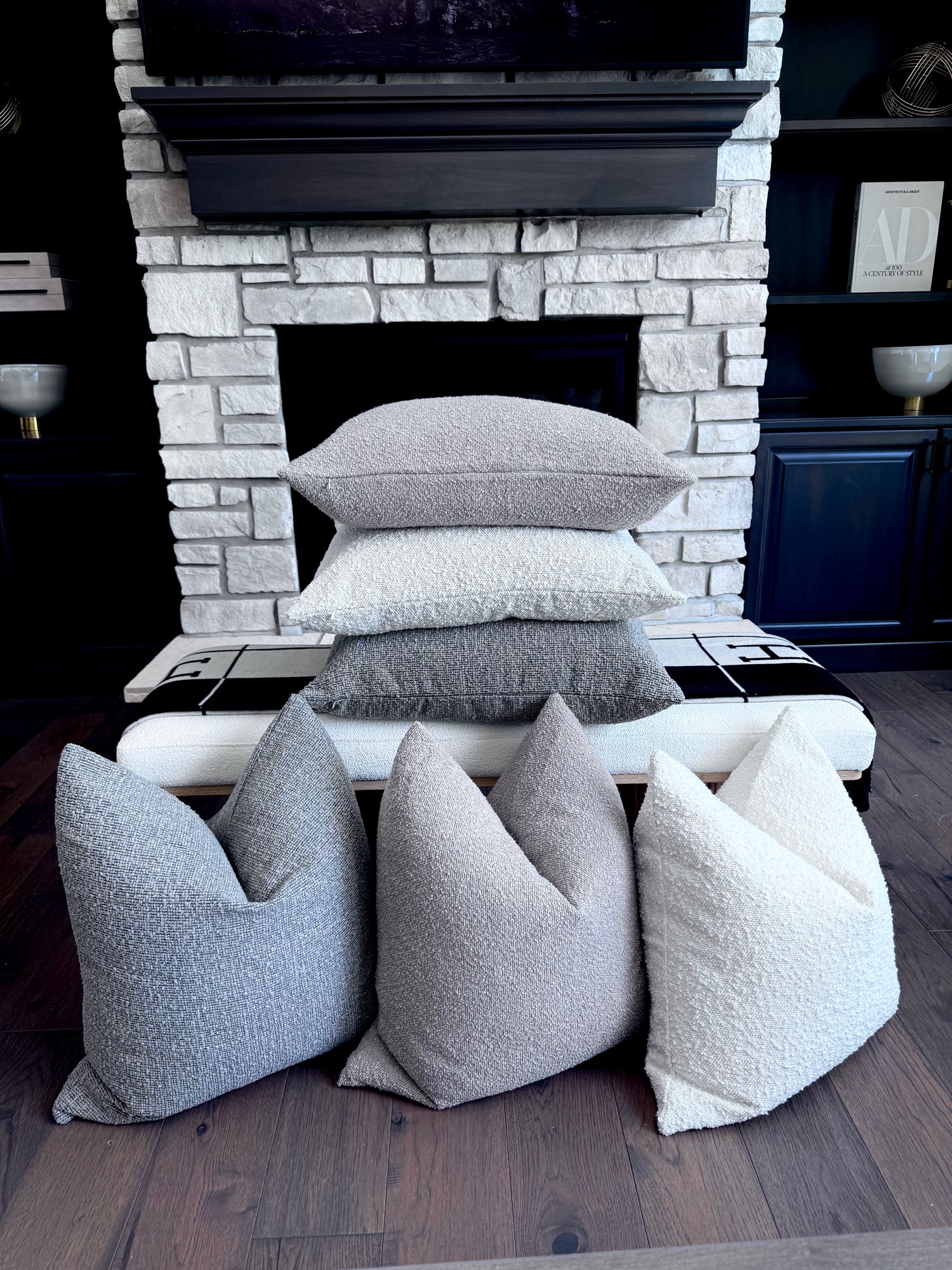 E.I.K. luxury cozy collection, three color textured throw pillows in various shades of gray and off-white are arranged on a bench and warm hardwood in front of a white stone fireplace with luxury touches.
