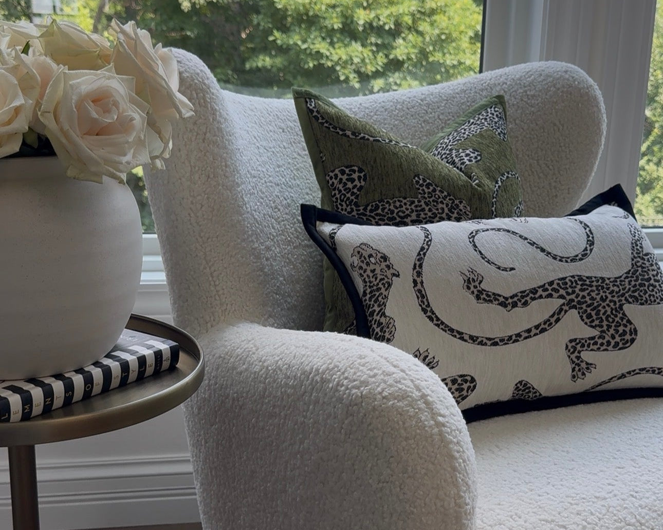 A cream bouclé armchair in a bright room by a window, styled with two E.I.K. Home Collection decorative throw pillows. A small side table with a white vase of white roses sits beside the chair.