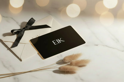 A black gift card with the white text 'EIK' lies on top of a cream envelope tied with a black ribbon, styled on a white marble surface with dried bunny tail grass and warm bokeh lights in the background.