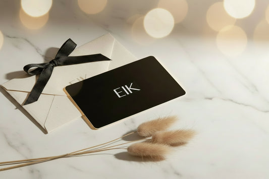 A black gift card with the white text 'EIK' lies on top of a cream envelope tied with a black ribbon, styled on a white marble surface with dried bunny tail grass and warm bokeh lights in the background.